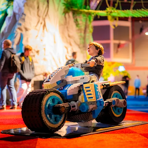 Een kind zit op een levensgrote motor gebouwd uit blauwe en gele LEGO® stenen