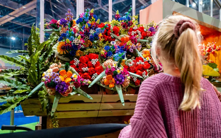 Een meisje bekijkt een kleurrijk boeket LEGO bloemen in een kruiwagen.