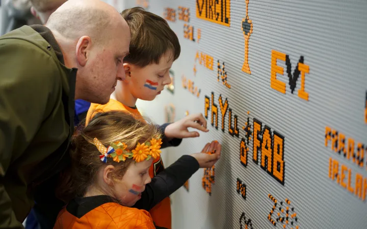 Een gezin in oranje voetbaltenue plaatst een steentje op een grote LEGO muur met namen van voetballers.
